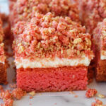 Three-layer pink cake bars with a white cream layer and a crumbly pink topping on a light surface, with crumbs scattered around