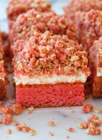 Three-layer pink cake bars with a white cream layer and a crumbly pink topping on a light surface, with crumbs scattered around