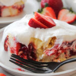 Sliced strawberry layer cake with white cream frosting and a strawberry on top, served on a plate with a black fork.